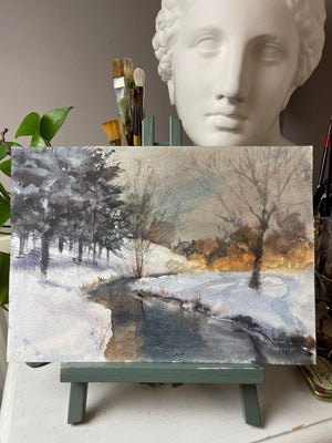 Original Winter River at Dusk watercolour on paper shown upright on an easel, with snowy banks and dark winding water.