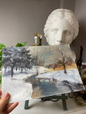 Winter River at Dusk original watercolour held at an angle, showing its paper surface and the snowy river landscape.